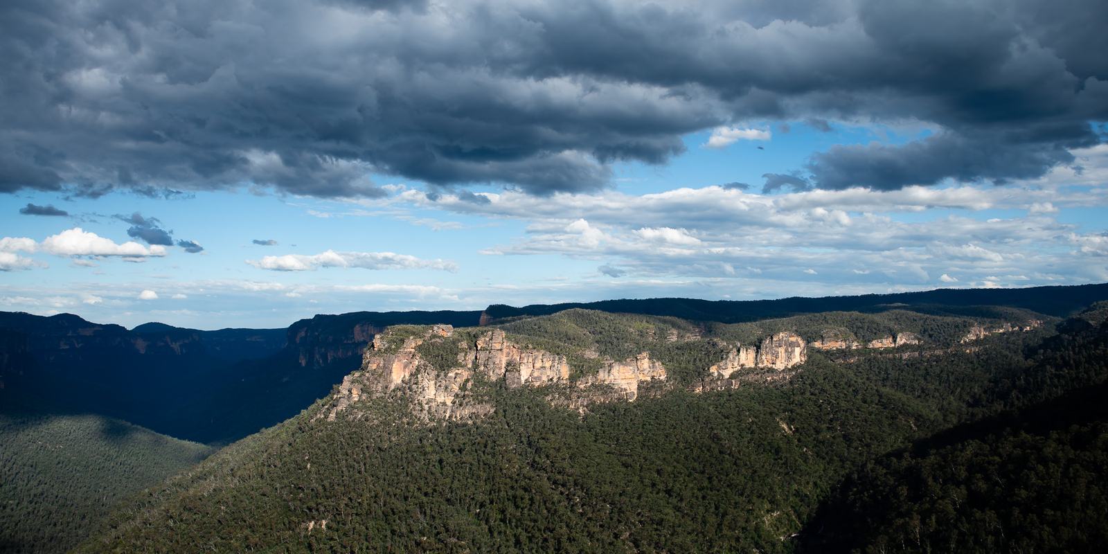 A long line of sandstone cliffs above forested slopes, lit from the side beneath heavy grey storm clouds.