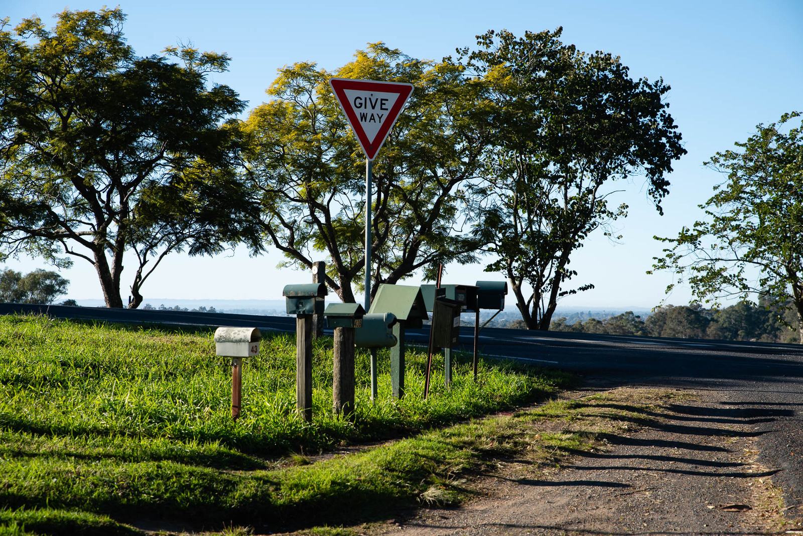 A cluster of rural letterboxes on timber posts beside a Give Way sign at a country road junction, with trees and a distant plain behind.